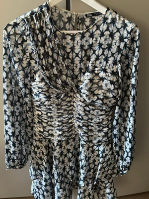 Blommig kortklänning från Zara - Säljer en snygg kortklänning från Zara med blommigt mönster i blått och vitt. Klänningen har långärmade ärmar, rynkade detaljer framtill och volang vid nederdelen. Materialet är lätt och luftigt, perfekt för vår och sommar.