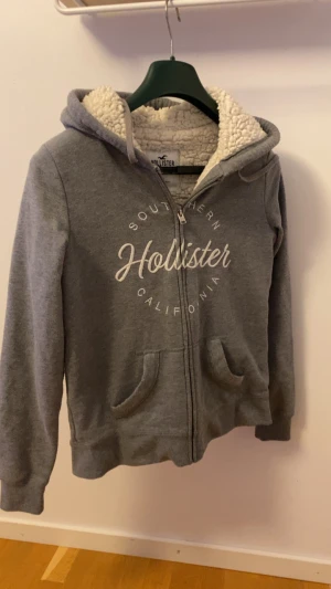 Grå hoodie med teddyfoder Hollister - Mysig grå hoodie från Hollister med fluffigt vitt teddyfoder på insidan. Tröjan har huva, dragkedja framtill och tryckt logga på bröstet. Perfekt för kalla dagar och chill hemma. Snygg och enkel att matcha med jeans eller mjukisbyxor.