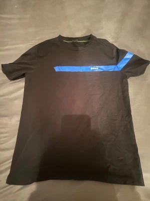 Svart BOSS t-shirt med blå detalj - Svart t-shirt från BOSS med en blå diagonal rand över bröstet och ärmen samt liten BOSS-logga. Klassisk rund hals och korta ärmar. Skön stretchig bomull som sitter snyggt och passar till många olika stilar.