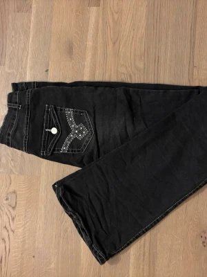 Svarta bootcut jeans med broderad ficka - Snygga svarta jeans med bootcut-snitt och kontrasterande vita sömmar. Bakfickan har ett broderat mönster och en knapp, vilket ger en unik detalj. Jeansen har klassisk femficksdesign och är tillverkade i jeansmaterial med dragkedja och knapp framtill.