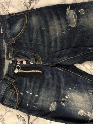 Dsquared2 blå jeans med dragkedja - Säljer ett par blå jeans från Dsquared2 med coola slitningar, färgstänk och en unik dragkedjedetalj framtill. Jeansen har klassisk femficksdesign, kontrastsömmar och Dsquared2-logga vid fickan. Perfekt för dig som gillar streetstyle och vill sticka ut.