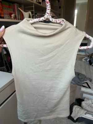 Beige ärmlös polotopp - Säljer en stilren beige polotopp utan ärmar. Toppen har en mjuk och stretchig känsla, perfekt för att matcha med jeans eller kjol. Enkel design med hög hals och rak passform.