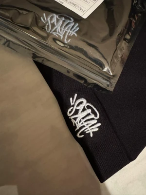 Svart mössa Syna - Svart mössa med vit broderad Syna-logga framtill. Enkel och stilren design med uppvikt kant. Perfekt för att hålla värmen och ge en cool touch till din outfit. Materialet känns mjukt och stretchigt, vilket gör den bekväm att bära. Helt ny. 