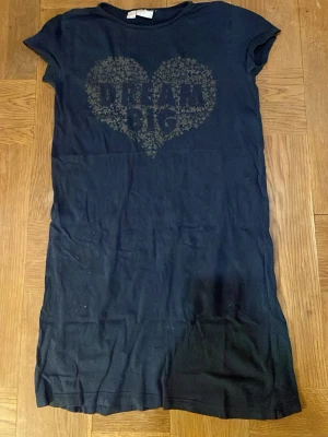 Mörkblå nattklänning med tryck från bpc - Snygg mörkblå nattklänning från bpc bonprix collection med rund hals och korta ärmar. Framsidan har ett stort hjärtformat tryck med texten 'DREAM BIG' och stjärnmönster. Klänningen är gjord i mjuk bomull och har en avslappnad passform. Väl använd men i bra skick.