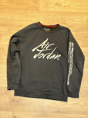 Svart Jordan sweatshirt med tryck - Svart sweatshirt från Jordan med stort Air Jordan-tryck på bröstet och vit text längs ena ärmen. Tröjan har rund hals, långa ärmar och röd detalj på insidan av kragen. Mjuk insida i fleece och klassisk loose passform.