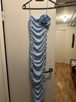 Ljusblå långklänning med rynk och blomma - Supersnygg ljusblå långklänning med smala axelband och rynkade detaljer längs hela klänningen. En stor tygblomma sitter på ena axeln för extra statement. Klänningen har figurnära passform. Materialet känns mjukt och stretchigt. Helt ny med prislappen kvar. Nypris 379 från Zara