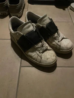 Valentino sneakers med svart detalj - Säljer ett par vita Valentino sneakers med bred svart panel över snörningen. Skorna har rund tå, platt sula och klassisk snörning. Tillverkade i skinn med robust gummisula där Valentino-loggan syns undertill. Perfekt för dig som gillar stilrena och lyxiga sneakers pris värda behöver bara tvättas