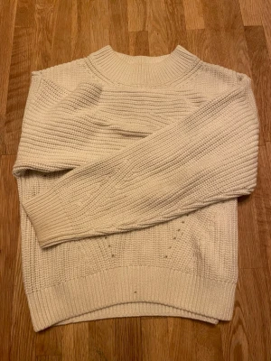 Vit stickad tröja från H&M - Säljer en vit stickad tröja från H&M med snygga detaljer och strukturstickning. Tröjan har en hög, ribbad hals och långa ärmar med breda muddar. Perfekt för dig som gillar en enkel men ändå cool look under höst och vinter. Tröjan har lite minifläckar vid muddarna men ingenting som man lägger märke till!