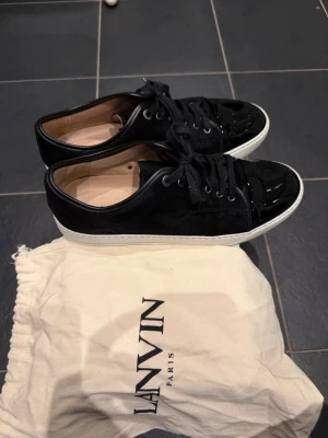 Svarta sneakers från Lanvin i mocka - Säljer ett par svarta Lanvin sneakers med ovandel i mocka och lackad tå. Skorna har vita sulor, svarta skosnören och metallfärgade öljetter. Klassisk och clean design med exklusiv känsla. Dustbag från Lanvin medföljer. Storlek UK 10 - 44 