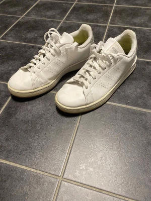Vita Adidas advantage sneakers  43⅓ - Fräscha vita Adidas advantage sneakers med klassisk låg profil och rund tå. Skorna har snörning, perforerade ränder på sidorna och grå detalj med Adidas-logga på hälen. Tillverkade i skinn med platt sula, perfekta för en clean och stilren look.