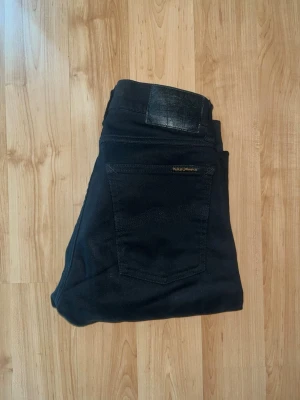 Nudie Jeans - Säljer ett par svarta jeans från Nudie Jeans med smal passform. Materialet är mjukt och stretchigt för extra komfort. Midja 33cm, längd 96cm. OBS jeansen har deffekt på både baksida lår och ficka baktill -se bild 6,7. JAG SKICKAR EJ FLER BILDER NÄR JAG BÄR JEANSEN! för bättre inblick i passformen rekommenderar jag att googla på modellnamnet. För storleksguide kolla måtten noggrant då jeansen alltid kan vara uppsydda heller krympta och jag ej tar emot returer :) s5,2