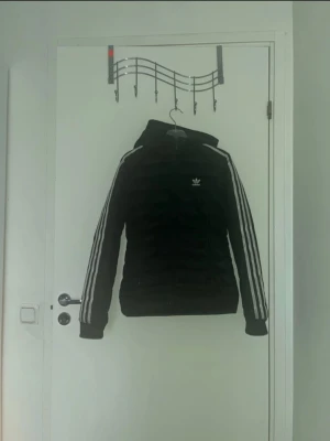 Svart pufferjacka från Adidas - Säljer en svart pufferjacka från Adidas med klassiska vita ränder längs ärmarna och Adidas-logga på bröstet. Jackan har dragkedja framtill, huva och quiltad design. Tillverkad i polyester och passar perfekt för kyligare dagar.