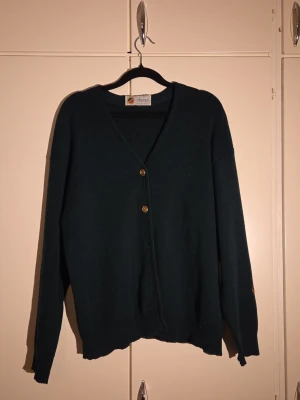 Busnel mörkgrön vintage kofta  i ull - Snygg mörkgrön kofta från Busnel, tillverkad i Frankrike av ren ull. Koftan har v-ringning, guldknappar framtill och långa ärmar. Klassisk och stilren design som passar perfekt till lager-på-lager. Busnel-logga på ärmen.