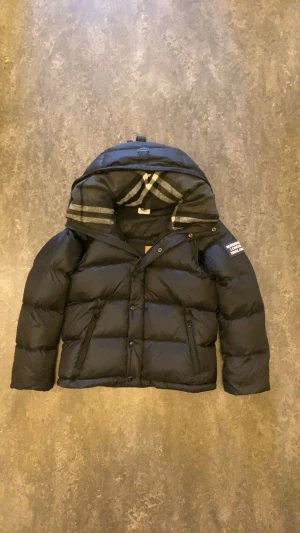 Svart dunjacka från Burberry med avtagbara ärmar - Svart pufferjacka från Burberry med avtagbara ärmar så du kan bära den som väst. Jackan har huva med klassiskt rutigt foder, dragkedja och knappar framtill samt två fickor med dragkedja. Perfekt för kalla dagar och riktigt snygg detalj med Burberry-logga på ärmen.