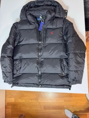 Svart pufferjacka RL - Svart pufferjacka från RL med huva, dragkedja och broderad röd logga på bröstet. Jackan har flera fickor med dragkedja och är tillverkad i polyester. Perfekt för kalla dagar och har en clean, modern look.