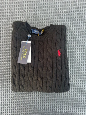 Svart kabelstickad tröja Polo Ralph Lauren - Svart kabelstickad tröja från Polo Ralph Lauren i storlek L men passar även M. Tröjan har rund hals, lång ärm och klassisk röd logobrodering på bröstet. Materialet är mjukt och stickat, perfekt för kyliga dagar. Snygg ribbad kant vid ärmslut och nederkant. Tröjan är i 100% bomull 