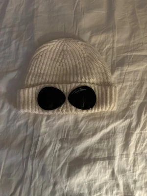 Vit ribbstickad mössa från C.P. Company - Säljer en vit ribbstickad mössa från C.P. Company med ikoniska svarta goggles på uppviket. Mössan är tillverkad i mjuk ull och har en clean, modern look som sticker ut. Perfekt för dig som vill ha något extra till vinterstilen.