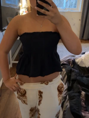 Svart bandeau tubtopp - Säljer en svart bandeau tubtopp med volangkant nedtill. Toppen är axelbandslös och har en croppad passform som sitter snyggt över bröstet. Perfekt för varma dagar och enkel att matcha med olika stilar.