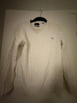 Vit kabelstickad tröja från GANT - Säljer en vit kabelstickad tröja från GANT i premium bomull. Tröjan har rund hals, ribbade muddar och ett litet broderat GANT-logo på bröstet. Perfekt för dig som gillar klassisk och stilren look!  Priset kan diskuteras!