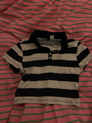  randig croppad polotopp - Säljer en croppad polotopp med breda mörkblåa och vita ränder. Toppen har korta ärmar, krage och tre knappar framtill. Materialet känns som bomull och toppen har en avslappnad passform som är perfekt till jeans eller kjol.🫰