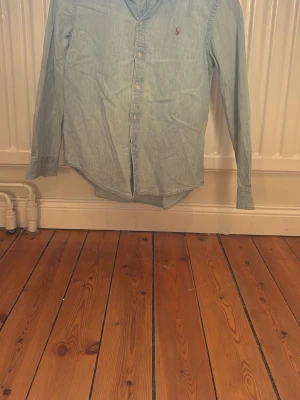 Ljusblå skjorta från Polo Ralph Lauren - Säljer en klassisk ljusblå skjorta från Polo Ralph Lauren med lång ärm och knappar framtill. Skjortan har en liten broderad logga på bröstet och är gjord i mjuk bomull. Perfekt för dig som gillar stilrena och tidlösa plagg.Några små fräckar