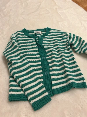 Randig grön kofta från Feelhoo XS/S - Säljer en stickad kofta från Feelhoo i storlek XS/S. Koftan har gröna och vita ränder, rund hals och knappar framtill. Materialet är stickat och ger en mysig känsla. Perfekt att slänga på sig när det är lite kyligt ute. 🥰 Aldrig använd.