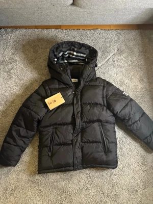 Svart pufferjacka från Burberry - Svart pufferjacka från Burberry med huva och snyggt rutigt foder. Jackan har två dragkedjefickor fram, knappar och en tydlig Burberry London England patch på ärmen. Perfekt för kalla dagar och har en riktigt schysst look.