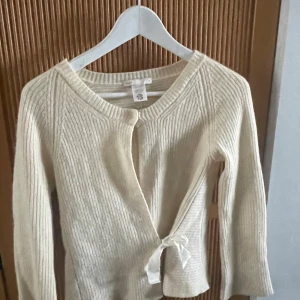 Vit kashmir omlottkofta från Filippa K - Säljer en elegant vit kofta från Filippa K i 100% kashmir. Koftan har en ribbad struktur och knytband framtill. Perfekt för en stilren look.