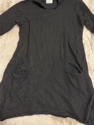 Svart klänning från Masai XL - Svart klänning från Masai Clothing Company i storlek XL. Modellen har lång ärm, rund hals och två fickor framtill. Materialet är mjukt och stickat, vilket gör den skön att bära. Enkel och stilren design som passar till många tillfällen.