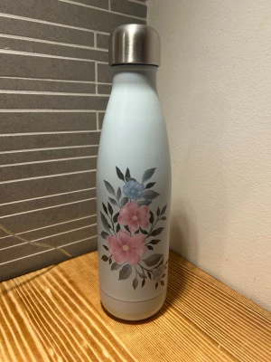 Blommig ljusblå termos vattenflaska från JobOut - Ljusblå termos vattenflaska från JobOut som rymmer 500 ml med blommigt mönster i rosa och blått framtill. Är i nytt skick och flaskan ska endast tvättas för hand. 