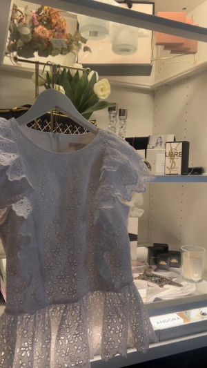 Ljusblå blus med volang och spets - Säljer en ljusblå blus med blommigt broderat spetsmönster och volangdetaljer på axlarna. Blusen har rund hals och är kortärmad med en söt peplumkant nedtill. Perfekt för dig som gillar romantisk och feminin stil.
