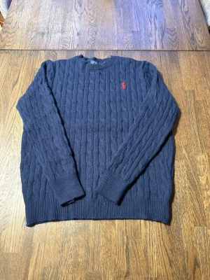 Mörkblå stickad tröja från Polo Ralph Lauren - Säljer en mörkblå stickad tröja från Polo Ralph Lauren med klassiskt kabelstickat mönster och röd broderad logga på bröstet. Tröjan har rund hals och långa ärmar med ribbade muddar vid ärmslut och nederkant. Perfekt för dig som gillar stilren och tidlös design.