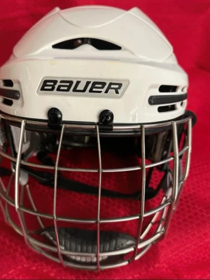 Bauer hockeyhjälm BHH9900L vit - Säljer en vit hockeyhjälm från Bauer, modell BHH9900L, med galler i metall och justerbar hakrem. Insidan har mjuk vaddering med EXPE foam för extra komfort och skydd. Ventilationshål på sidorna och bak för bra luftflöde. Passar dig som vill ha säkerhet och stil på isen.