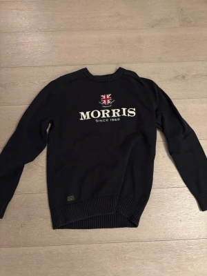 Svart stickad tröja från Morris - Svart stickad tröja från Morris Gents i storlek M. Tröjan har ett broderat Morris-tryck och brittisk flagga på bröstet, samt ribbade muddar vid ärmslut, hals och nederkant. Perfekt för dig som gillar klassisk stil med snygga detaljer.