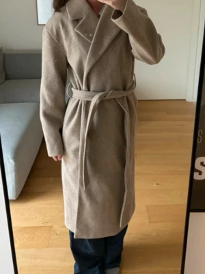 Beige kappa med bälte från Gina Tricot - Säljer en snygg beige kappa från Gina Tricot i storlek XS. Modellen har klassisk krage, knytbälte i midjan och slits bak. Kappan är fodrad och har långärmade ärmar, perfekt för dig som gillar stilrena och tidlösa plagg.