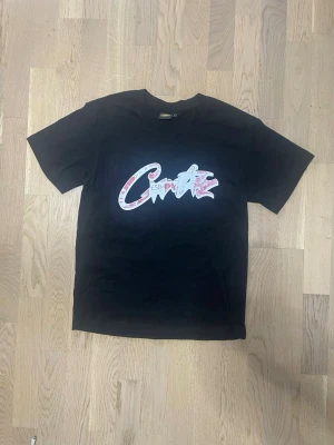 Svart Corteiz t-shirt med tryck - Svart t-shirt med fet grafisk print i vitt och rött på bröstet där det står 'Corteiz' i graffiti-stil. Rund hals och korta ärmar. Materialet är mjuk bomull som känns skönt mot huden. Perfekt för dig som gillar streetwear och vill sticka ut med coola detaljer. T shirten är storlek S men M fungerar bra också! Den är som ny, så hör av dig vid intresse. Pris kan diskuteras!