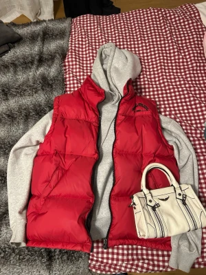 Röd dunväst med dragkedja - Röd dunväst från S.Oliver Athletic med svart broderad logga på bröstet. Västen har hög krage, två fickor och stängs med dragkedja. Perfekt att slänga över en hoodie för en chill och sportig look.