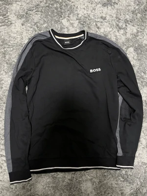Svart långärmad tröja från BOSS - Säljer en svart långärmad tröja från BOSS med vit logga på bröstet och gråa detaljer längs ärmarna. Tröjan har rund halsringning med vit kant och är tillverkad i mjuk bomull. Perfekt för en clean och stilren look.