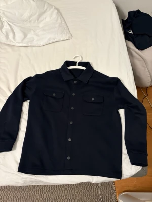 Mörkblå overshirt med knappar - Snygg mörkblå overshirt med klassisk krage och två stora bröstfickor med lock och knappar. Jackan har långa ärmar och stängs med knappar framtill. Perfekt för lager-på-lager och enkel att styla till olika outfits.