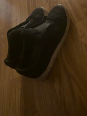 Svarta arigato skor  - Svarta arigato skor med vit sula. Skorna har en enkel och stilren design utan snörning, vilket gör dem lätta att ta på och av. Ovandelen ser ut att vara i textil eller syntetmaterial och sulan är platt. Perfekta för en avslappnad och bekväm stil.