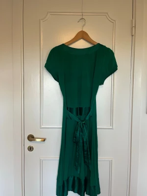 Grön klänning - Säljer en smaragdgrön långklänning från Paris Kate & I i storlek M. Klänningen har korta ärmar, rundad urringning och ett snyggt band i midjan som knyts bak. Materialet känns mjukt och faller fint, med en diskret glansig kant längst ner.