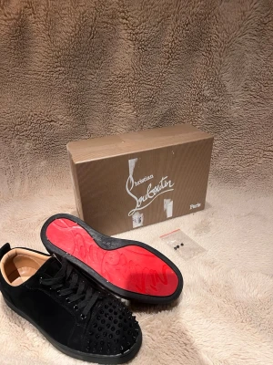 Christian Louboutin svarta sneakers med nitar - Säljer ett par svarta sneakers från Christian Louboutin i mocka med ikoniska röda sulor och svarta nitar på tån. Skorna har snörning och en rund tå, samt lyxig läderinsida. Perfekta för dig som vill sticka ut med edgy detaljer och exklusiv känsla.