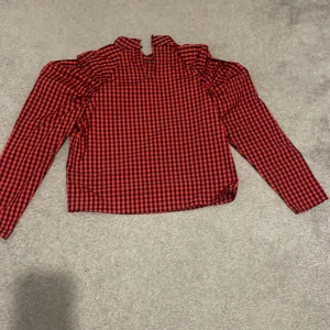 Rutig blus med volanger  -   Röd-svart rutig blus från H&M i storlek 164. Långärmad modell med fina volangdetaljer vid axlarna. Bekväm passform och mjukt material – perfekt både till vardag och fest. I mycket fint skick.