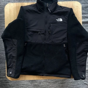 Svart Denali fleece från The North Face - Svart fleecejacka från The North Face i herrmodell, storlek XS. Jackan har hög krage, flera fickor med dragkedja och bröstficka med spänne. Materialmix av fleece och syntet, perfekt för kyliga dagar. Snyggt broderad logga på bröstet och ryggen. Bra omhändertagen. Originalpris: 2200kr