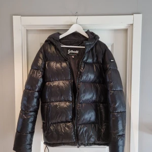 Svart pufferjacka från Schott  - Schysst svart pufferjacka från Schott med glansig finish och huva. Jackan har dragkedja framtill, två sidofickor med dragkedja och Schott-logga på ärmen. Perfekt för kalla dagar och har en riktigt modern look. Knappt använd köpte förra vintern men var inget för mig då jag ville ha en matt. Ny pris 2699!