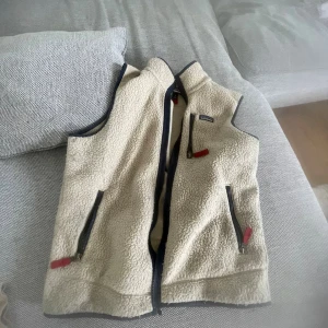 Beige fleeceväst från Patagonia - Säljer en beige fleeceväst från Patagonia med svarta detaljer och dragkedjor. Västen har tre fickor med dragkedja och röda zip-pullers. Perfekt för lager-på-lager och chillare dagar. Klassisk och enkel design med hög krage.