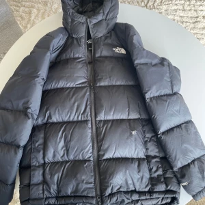 Svart dunjacka från The North Face - Svart dunjacka från The North Face med klassisk pufferstil och huva. Jackan har dragkedja framtill, två sidofickor och logga på bröstet.  finns ett litet hål, se bild (går att laga lätt) Stygn har släppt i nederkant , se bild.                           Strl S  500:- 