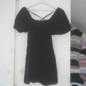 Svart kort klänning med puffärm  - Supersöt svart kort klänning från H&M Divided med puffiga ärmar, smockad rygg och snygga knytband i ryggen. Perfekt för dig som gillar trendiga detaljer och vill sticka ut.