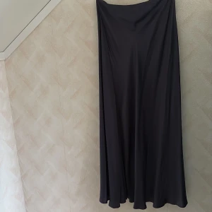 Mörkblå långkjol från Zara - Säljer en stilren mörkblå långkjol från Zara i flowigt material. Kjolen har en lätt glansig yta och stängs med dragkedja där bak. Jag har aldrig använt den samt så finns prislappen kvar.