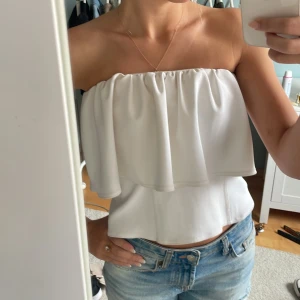 Vit axelbandslös tubtopp med volang - Säljer en superfin vit axelbandslös tubtopp med volangdetalj. Toppen har bandeau-modell och är helt ärmlös, perfekt till jeans eller kjol. Materialet känns mjukt och har ett snyggt fall. Enkel och stilren design som ger en fräsch look. 